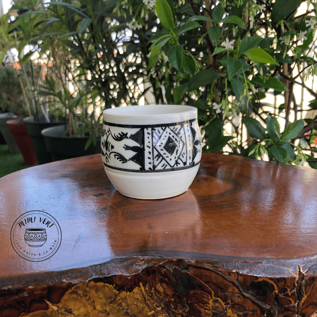 Pot en céramique fait à la main avec des motifs inspirés de l’artisanat marocain, noir et blanc, posé sur une table en bois avec des plantes vertes en arrière-plan.