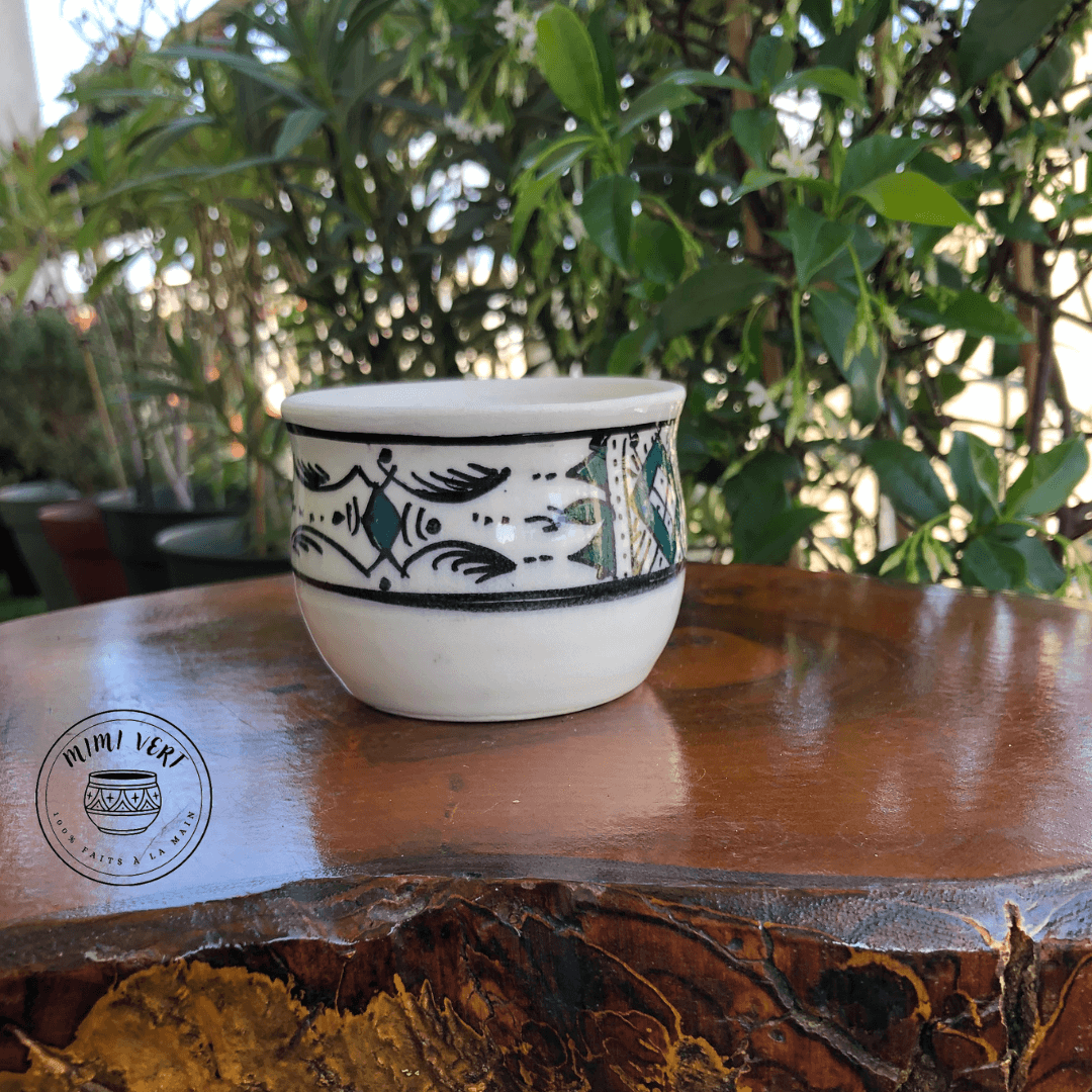 Pot en céramique fait à la main avec des motifs inspirés de l’artisanat marocain, blanc, noir et vert, posé sur une table en bois avec des plantes vertes en arrière-plan.