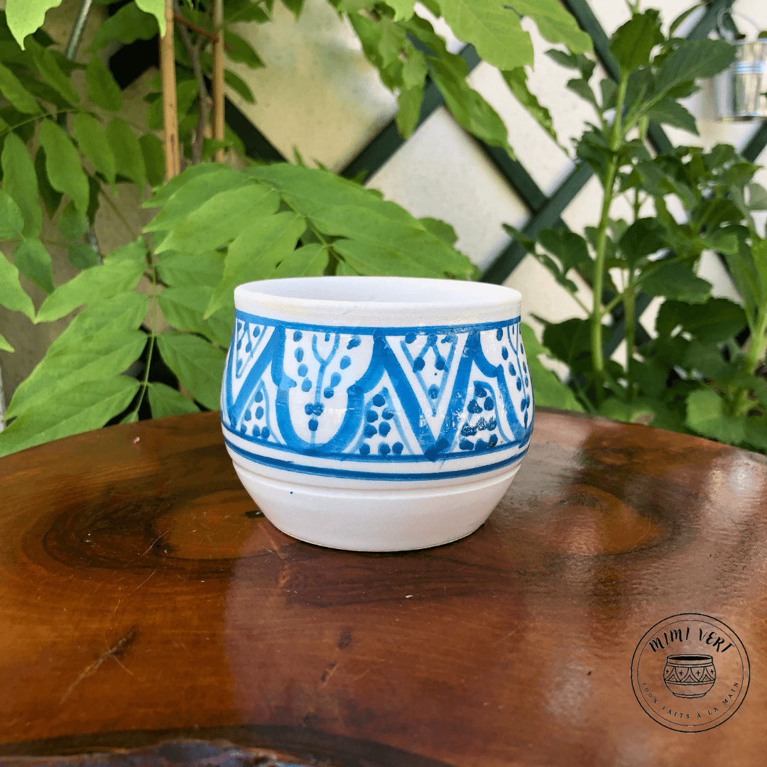 Pot en céramique fait à la main avec des motifs inspirés de l’artisanat marocain, bleu et blanc, posé sur une table en bois avec des plantes vertes en arrière-plan.