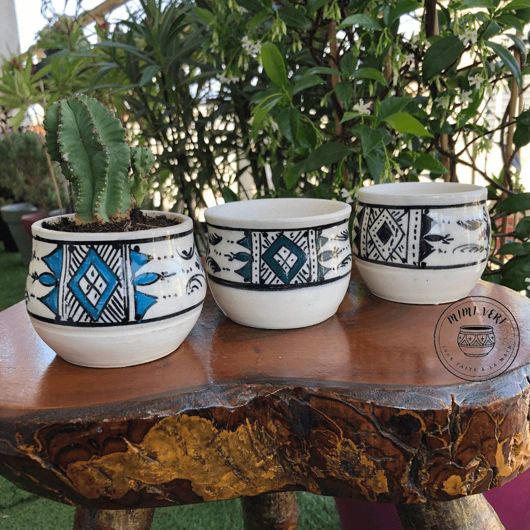 Trois pots en céramique faits à la main avec des motifs inspirés de l’artisanat marocain, posés sur une table en bois avec des plantes vertes en arrière-plan, dont un contenant un cactus.