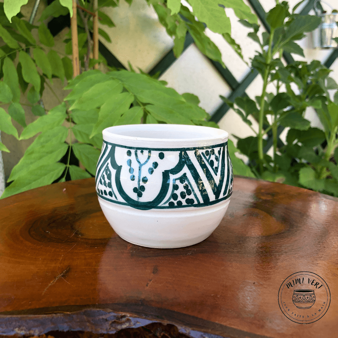 Pot en céramique fait à la main avec des motifs inspirés de l’artisanat marocain, blanc et vert, posé sur une table en bois avec des plantes vertes en arrière-plan.