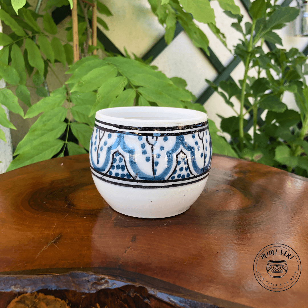 Pot en céramique fait à la main avec des motifs inspirés de l’artisanat marocain, bleu noir et blanc, posé sur une table en bois avec des plantes vertes en arrière-plan.