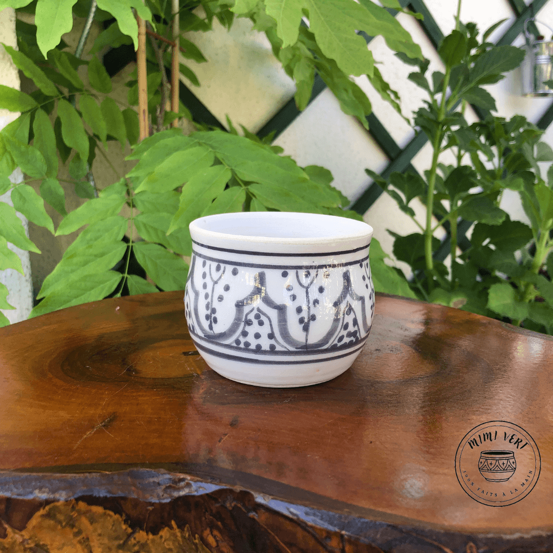 Pot en céramique fait à la main avec des motifs inspirés de l’artisanat marocain, gris et blanc, posé sur une table en bois avec des plantes vertes en arrière-plan.