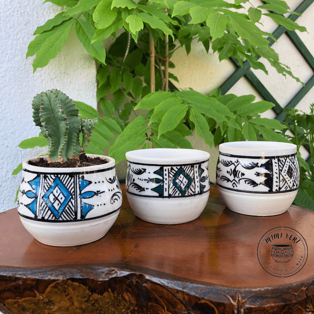 Trois pots en céramique faits à la main avec des motifs inspirés de l’artisanat marocain, posés sur une table en bois avec des plantes vertes en arrière-plan, dont un contenant un cactus.