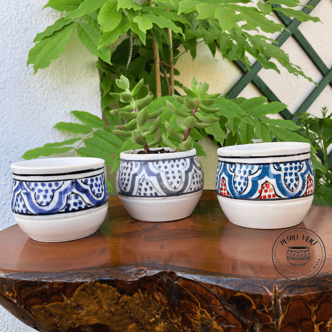 Trois pots en céramique faits à la main avec des motifs inspirés de l’artisanat marocain, posés sur une table en bois avec des plantes vertes en arrière-plan, dont un contenant une plante grasse.