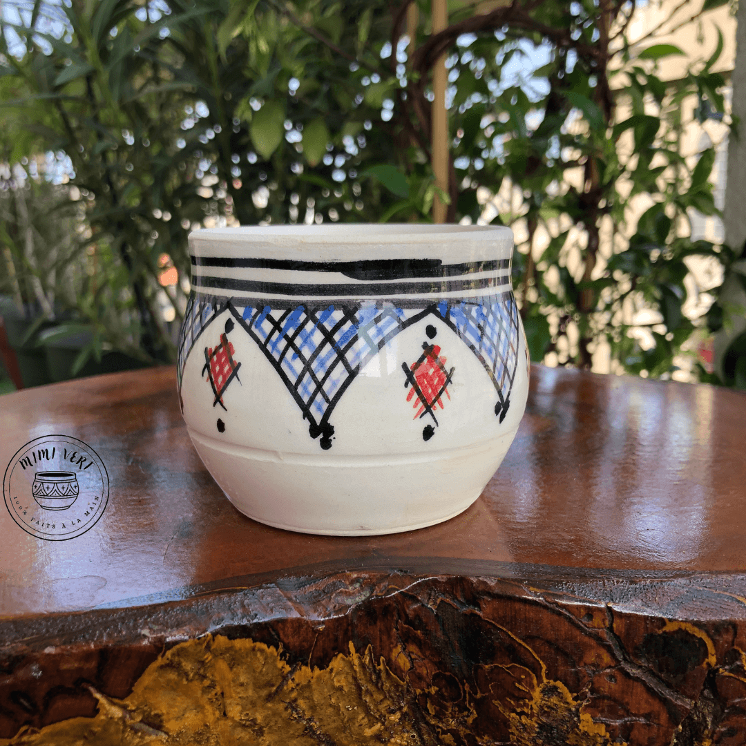Pot en céramique fait à la main avec des motifs inspirés de l’artisanat marocain, blanc noir rouge et bleu, posé sur une table en bois avec des plantes vertes en arrière-plan.