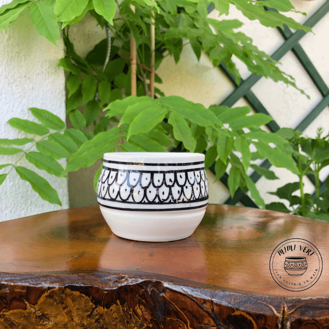Pot en céramique fait à la main avec des motifs inspirés de l’artisanat marocain, noir et blanc, contenant une plante grasse, posé sur une table en bois avec des plantes vertes en arrière-plan.