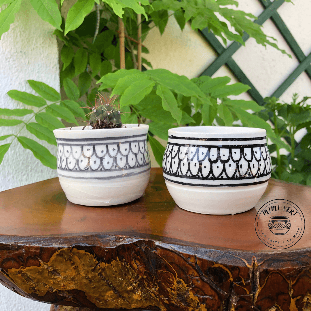 Deux pots en céramique faits à la main avec des motifs inspirés de l’artisanat marocain, posés sur une table en bois avec des plantes vertes en arrière-plan, dont un contenant un cactus.