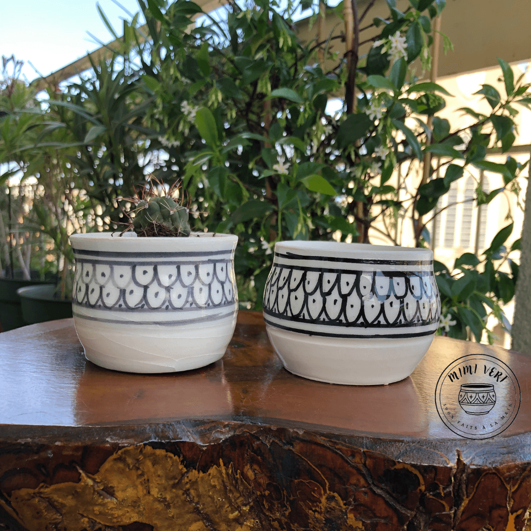 Deux pots en céramique faits à la main avec des motifs inspirés de l’artisanat marocain, posés sur une table en bois avec des plantes vertes en arrière-plan, dont un contenant un cactus.