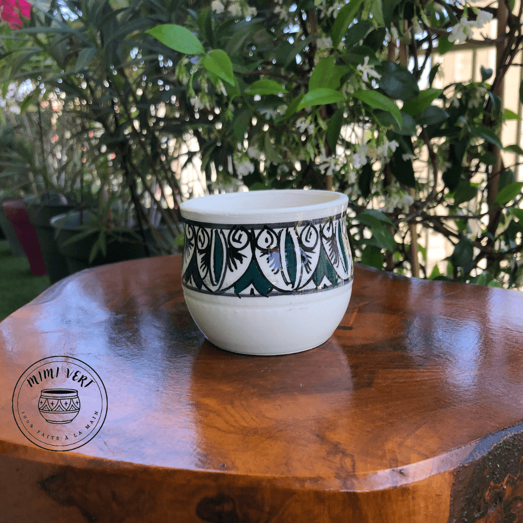 Pot en céramique fait à la main avec des motifs inspirés de l’artisanat marocain, blanc noir et vert, posé sur une table en bois avec des plantes vertes en arrière-plan.