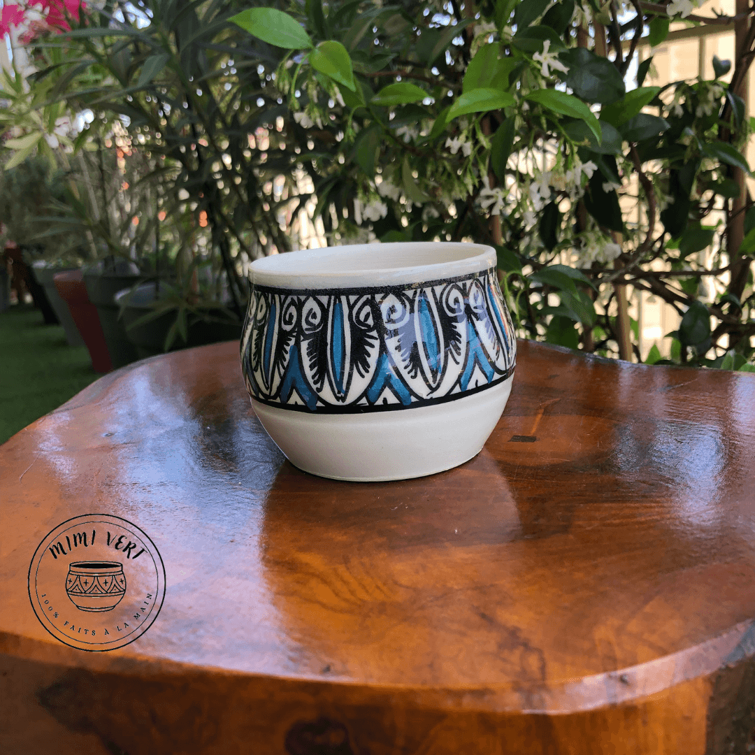 Pot en céramique fait à la main avec des motifs inspirés de l’artisanat marocain, bleu noir et blanc, posé sur une table en bois avec des plantes vertes en arrière-plan.