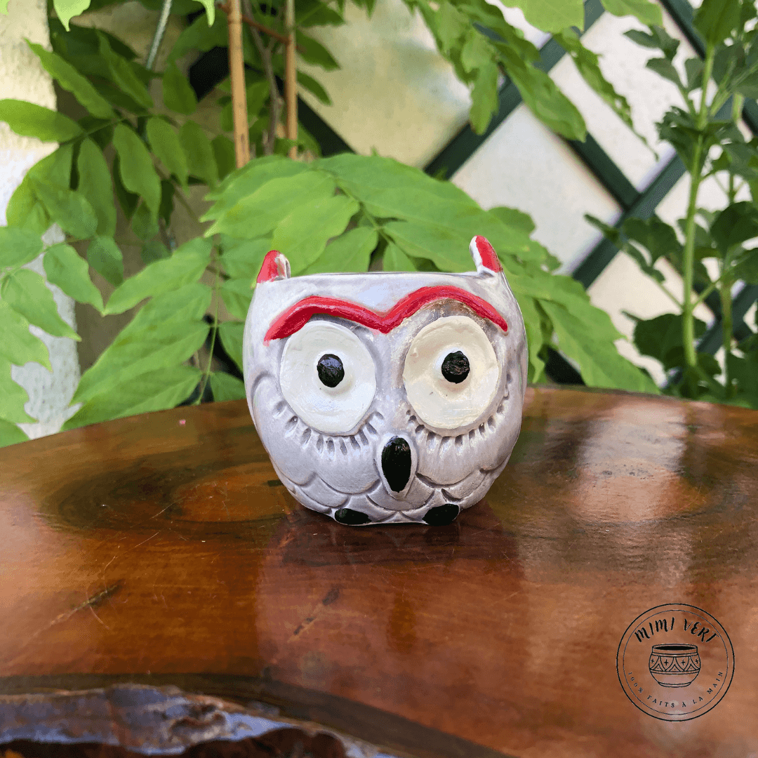 Petit pot en forme de hibou, gris clair, posé sur une table en bois avec des plantes vertes en arrière-plan.
