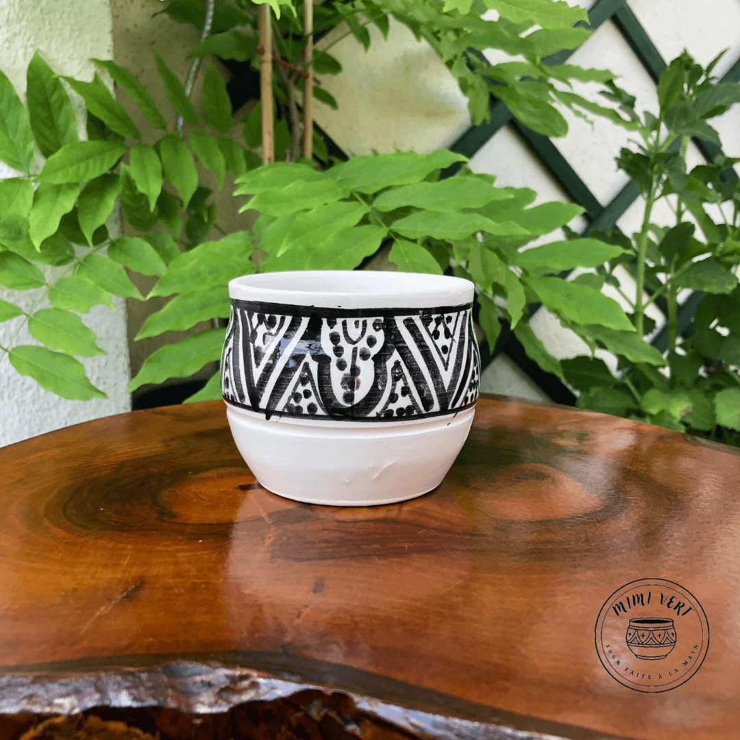 Pot en céramique fait à la main avec des motifs inspirés de l’artisanat marocain, blanc et noir, contenant un cactus, posé sur une table en bois avec des plantes vertes en arrière-plan.