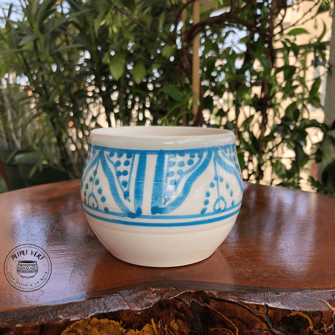 Pot en céramique fait à la main avec des motifs inspirés de l’artisanat marocain, bleu et blanc, posé sur une table en bois avec des plantes vertes en arrière-plan.