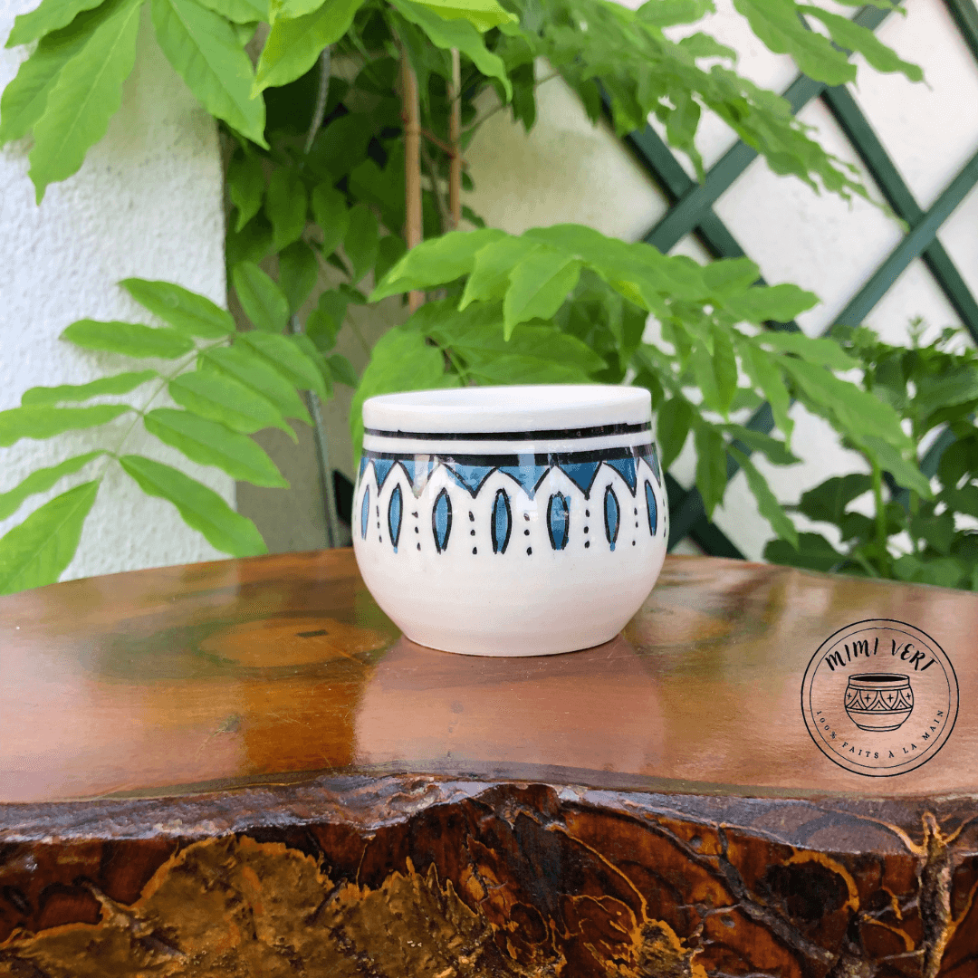 Pot en céramique fait à la main avec des motifs inspirés de l’artisanat marocain, bleu et blanc, posé sur une table en bois avec des plantes vertes en arrière-plan.