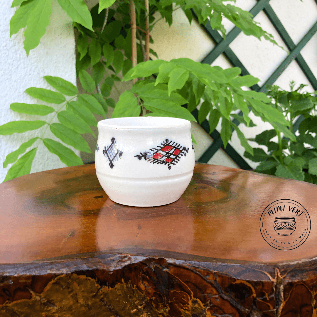 Pot en céramique fait à la main avec des motifs inspirés de l’artisanat marocain, blanc bleu et rouge, posé sur une table en bois avec des plantes vertes en arrière-plan.