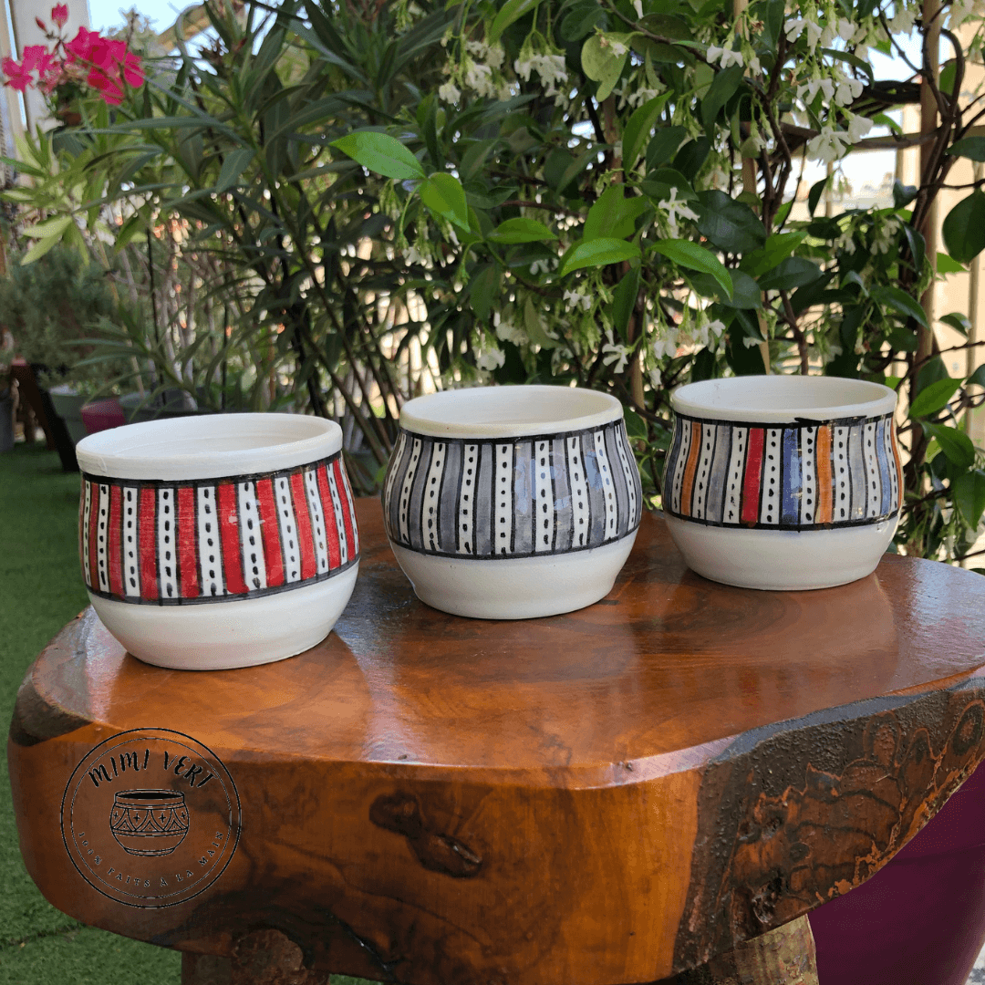 Trois pots en céramique faits à la main avec des motifs inspirés de l’artisanat marocain, sans plantes, posés sur une table en bois avec des plantes vertes en arrière-plan.