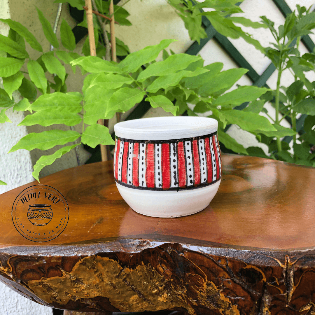 Pot en céramique fait à la main avec des motifs inspirés de l’artisanat marocain, blanc et rouge, posé sur une table en bois avec des plantes vertes en arrière-plan.