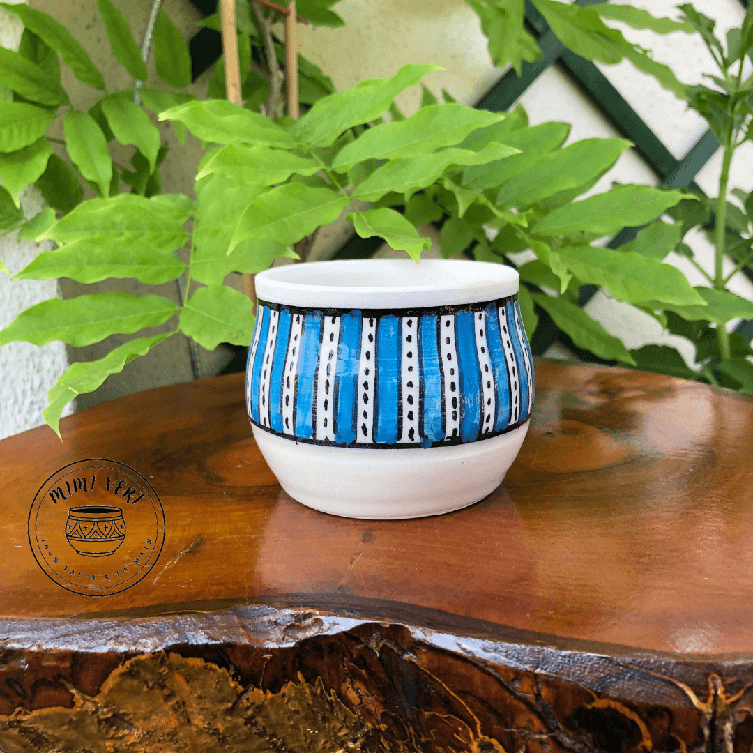 Pot en céramique fait à la main avec des motifs inspirés de l’artisanat marocain, blanc et bleu, posé sur une table en bois avec des plantes vertes en arrière-plan.