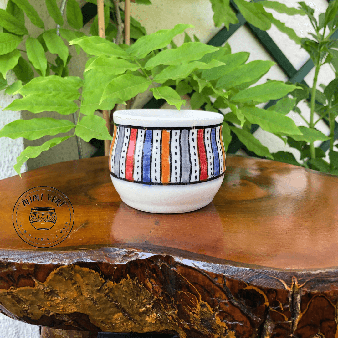 Pot en céramique fait à la main avec des motifs inspirés de l’artisanat marocain, blanc et multicolore, posé sur une table en bois avec des plantes vertes en arrière-plan.
