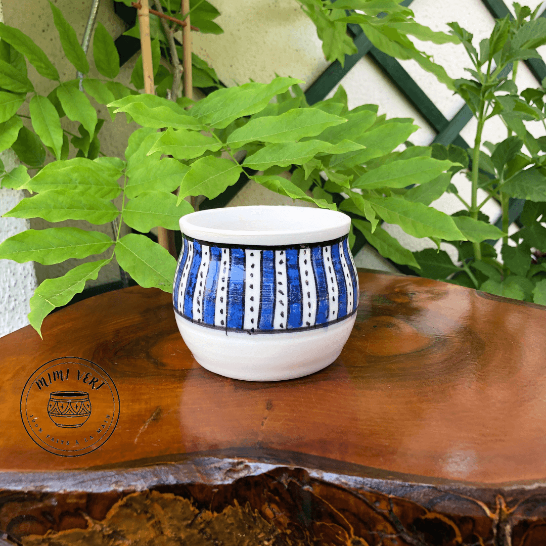 Pot en céramique fait à la main avec des motifs inspirés de l’artisanat marocain, blanc et bleu, posé sur une table en bois avec des plantes vertes en arrière-plan.