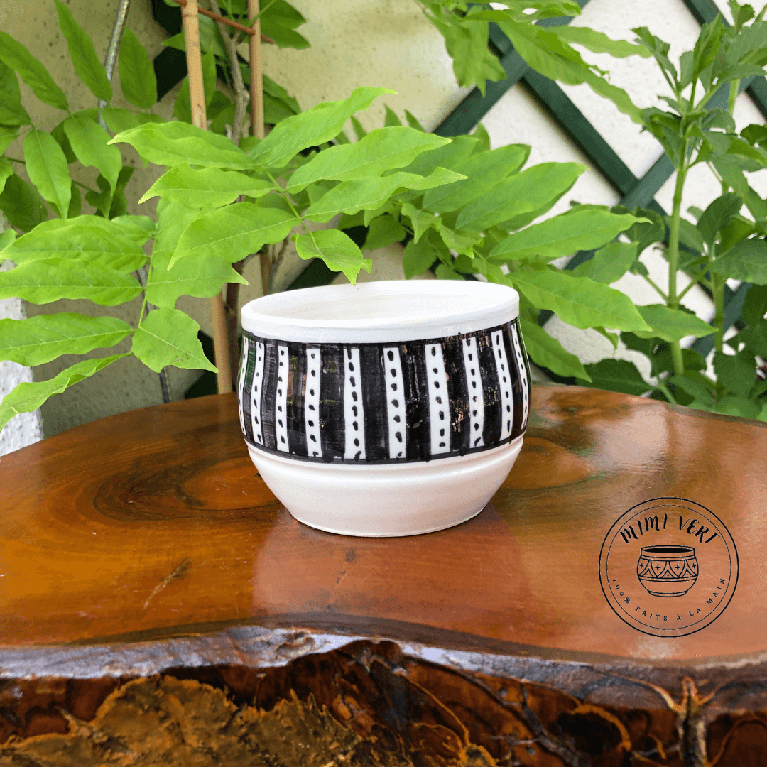 Pot en céramique fait à la main avec des motifs inspirés de l’artisanat marocain, blanc et noir, posé sur une table en bois avec des plantes vertes en arrière-plan.