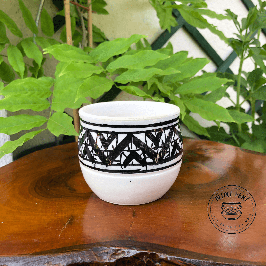 Pot en céramique fait à la main avec des motifs inspirés de l’artisanat marocain noir et blanc, posé sur une table en bois avec des plantes vertes en arrière-plan.