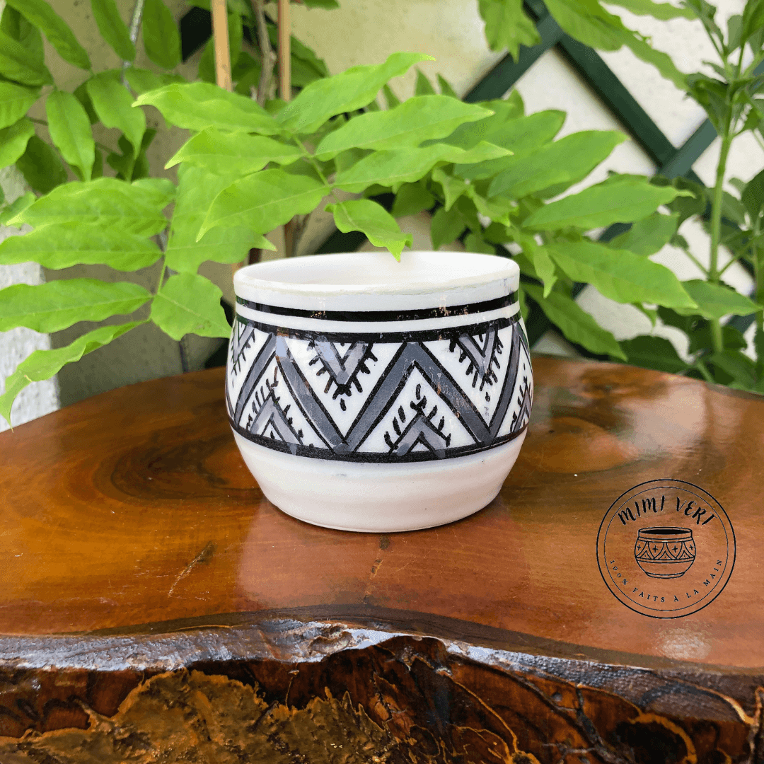 Pot en céramique fait à la main avec des motifs inspirés de l’artisanat marocain gris, noir et blanc, posé sur une table en bois avec des plantes vertes en arrière-plan.