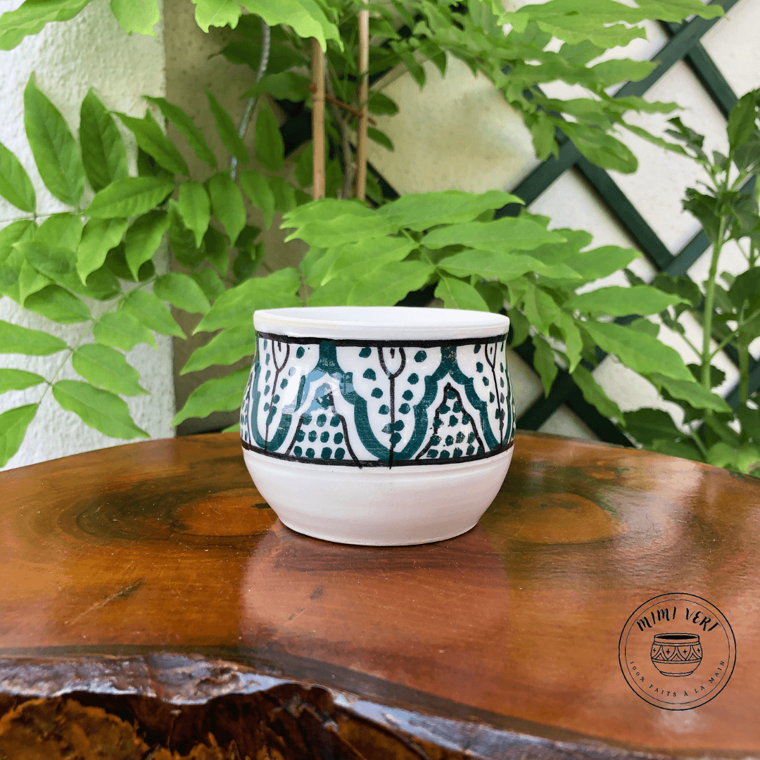 Pot en céramique fait à la main avec des motifs inspirés de l’artisanat marocain, blanc vert et noir, contenant un cactus, posé sur une table en bois avec des plantes vertes en arrière-plan.