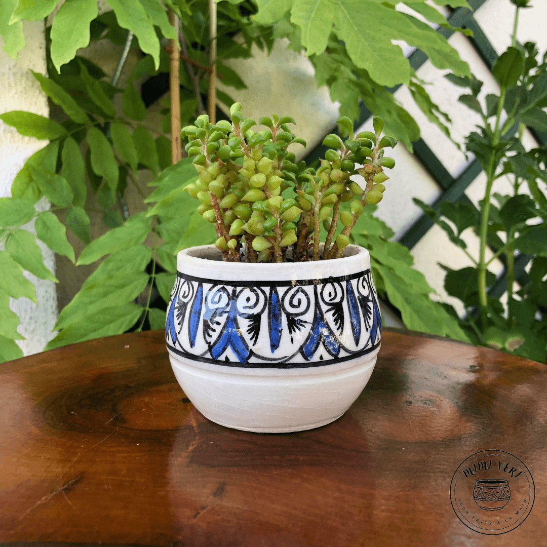 Pot en céramique fait à la main avec des motifs inspirés de l’artisanat marocain, bleu noir et blanc, posé sur une table en bois avec des plantes vertes en arrière-plan.