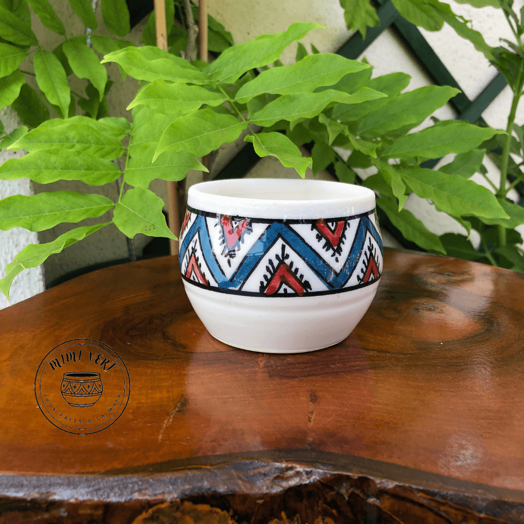 Pot en céramique fait à la main avec des motifs inspirés de l’artisanat marocain turquoise, rouge, blanc, posé sur une table en bois avec des plantes vertes en arrière-plan.