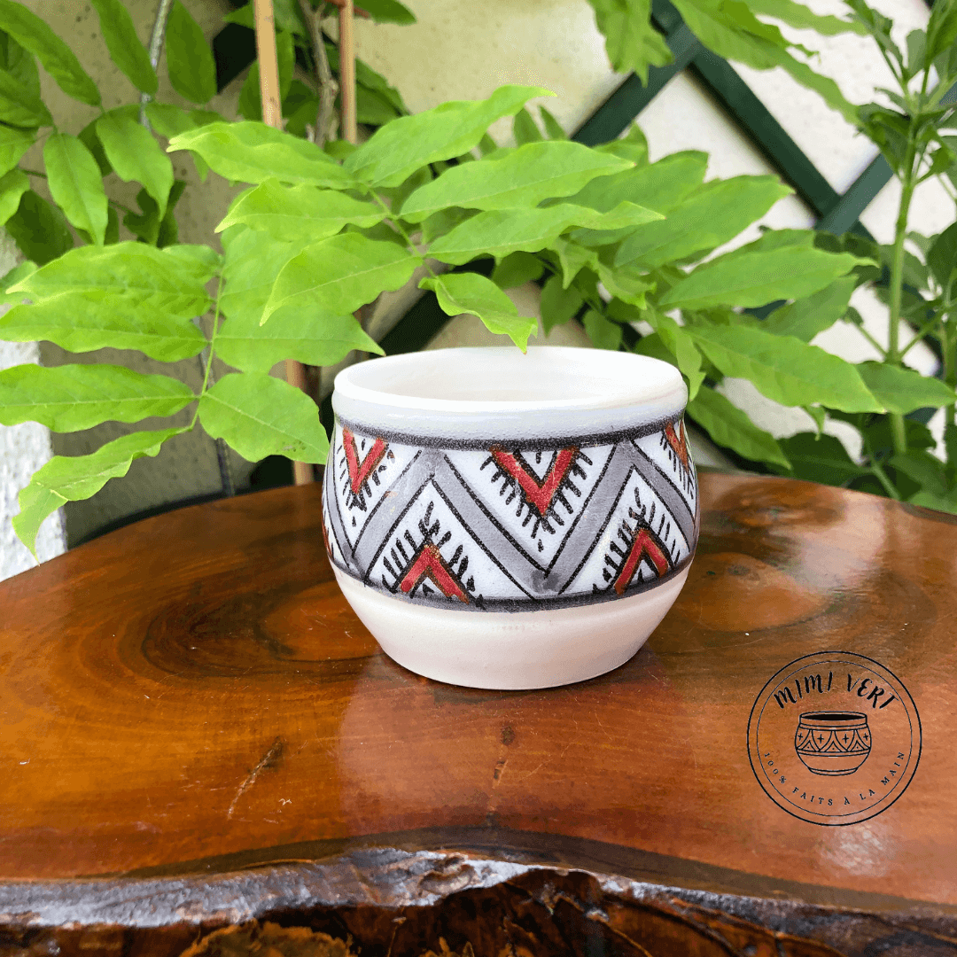 Pot en céramique fait à la main avec des motifs inspirés de l’artisanat marocain gris, rouge, blanc, posé sur une table en bois avec des plantes vertes en arrière-plan.