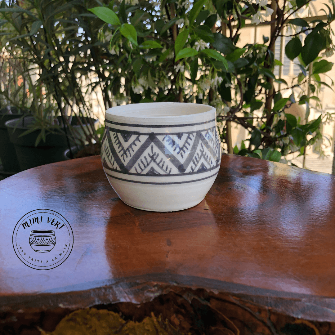 Pot en céramique fait à la main avec des motifs inspirés de l’artisanat marocain gris et blanc posé sur une table en bois avec des plantes vertes en arrière-plan.