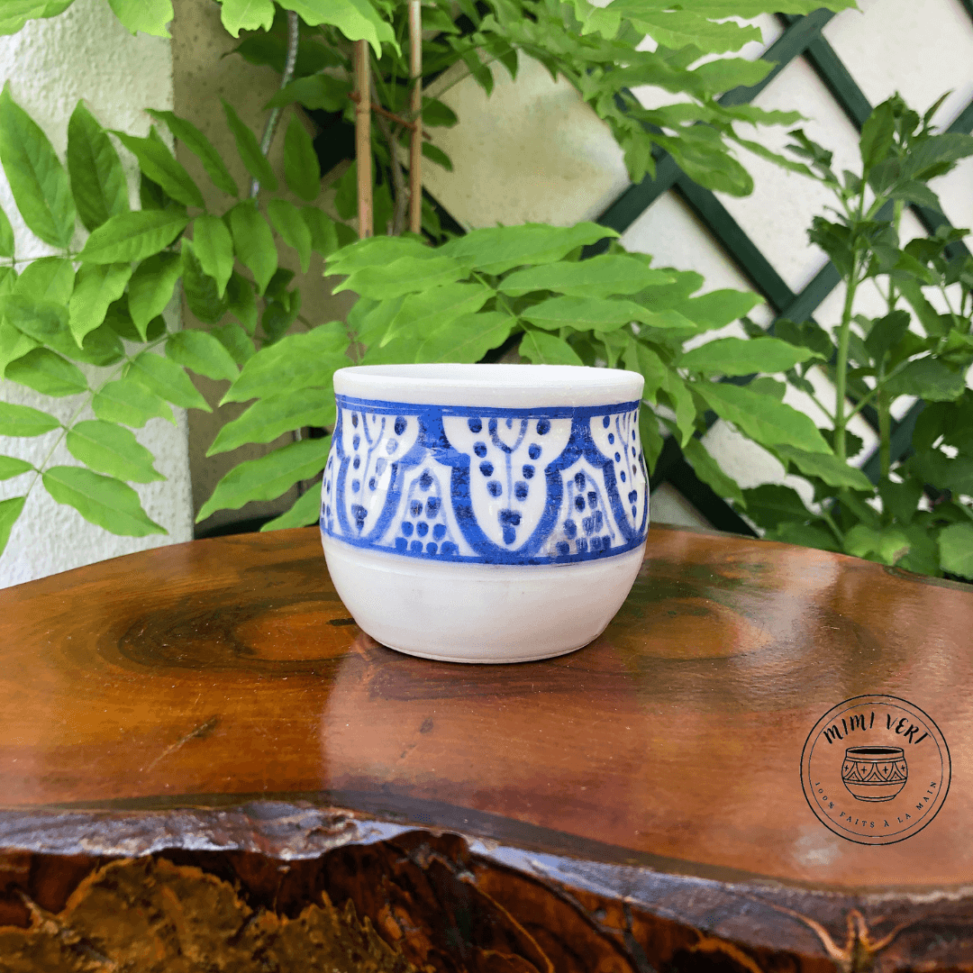 Pot en céramique fait à la main avec des motifs inspirés de l’artisanat marocain, blanc et bleu, contenant un cactus, posé sur une table en bois avec des plantes vertes en arrière-plan.