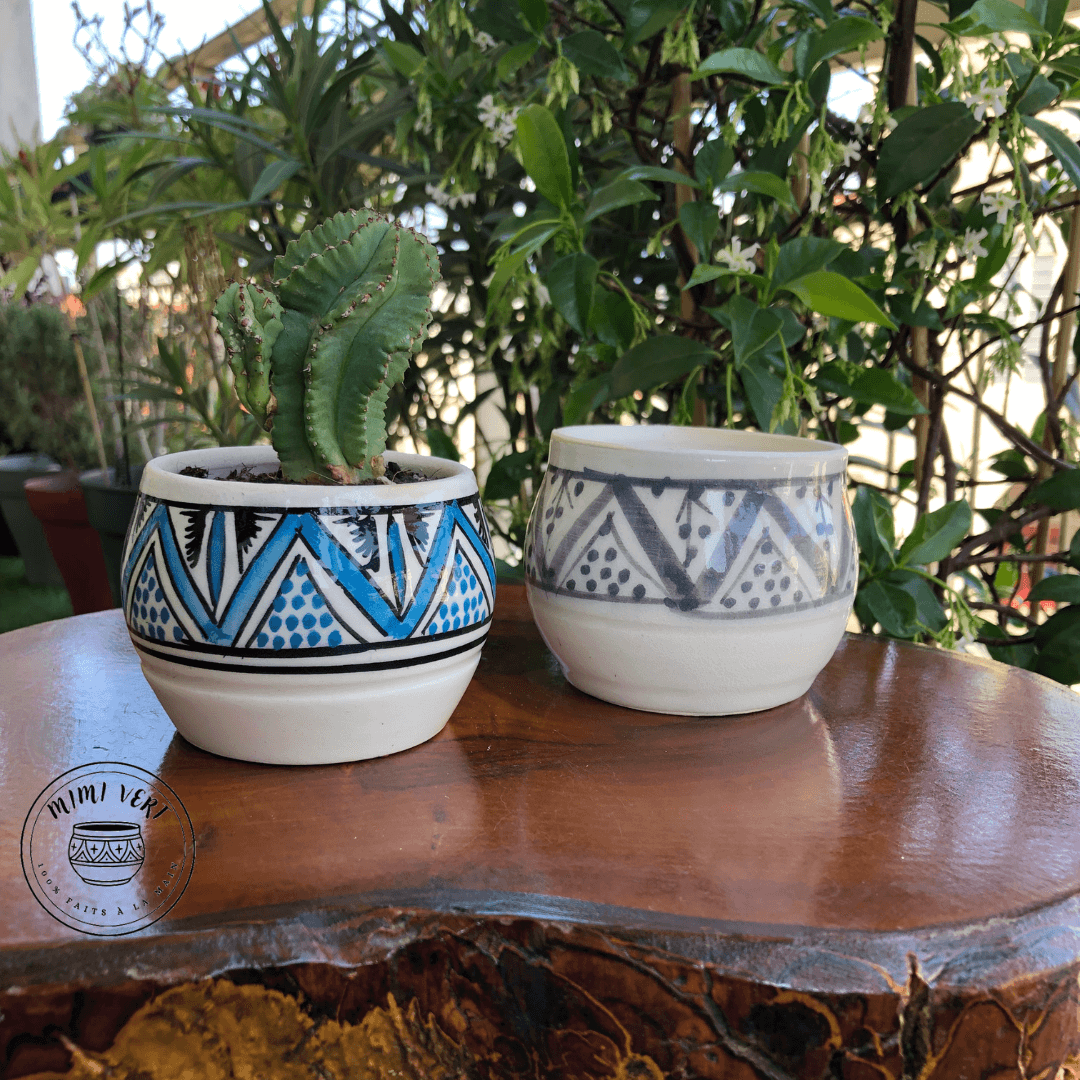 Deux pots en céramique faits à la main avec des motifs inspirés de l’artisanat marocain, posés sur une table en bois avec des plantes vertes en arrière-plan, dont un contenant un cactus.
