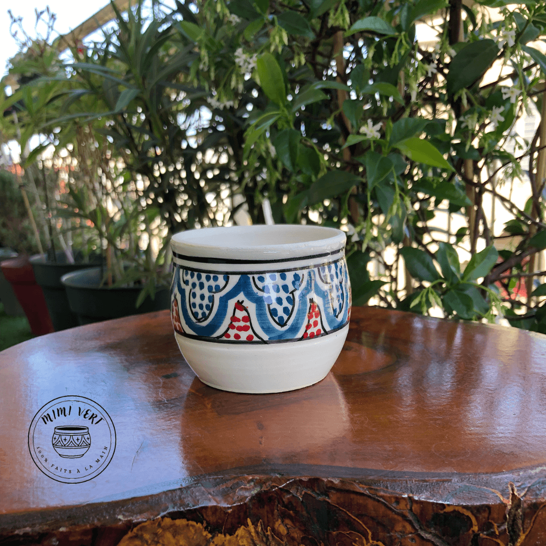 Pot en céramique fait à la main avec des motifs inspirés de l’artisanat marocain, bleu rouge et blanc, posé sur une table en bois avec des plantes vertes en arrière-plan.