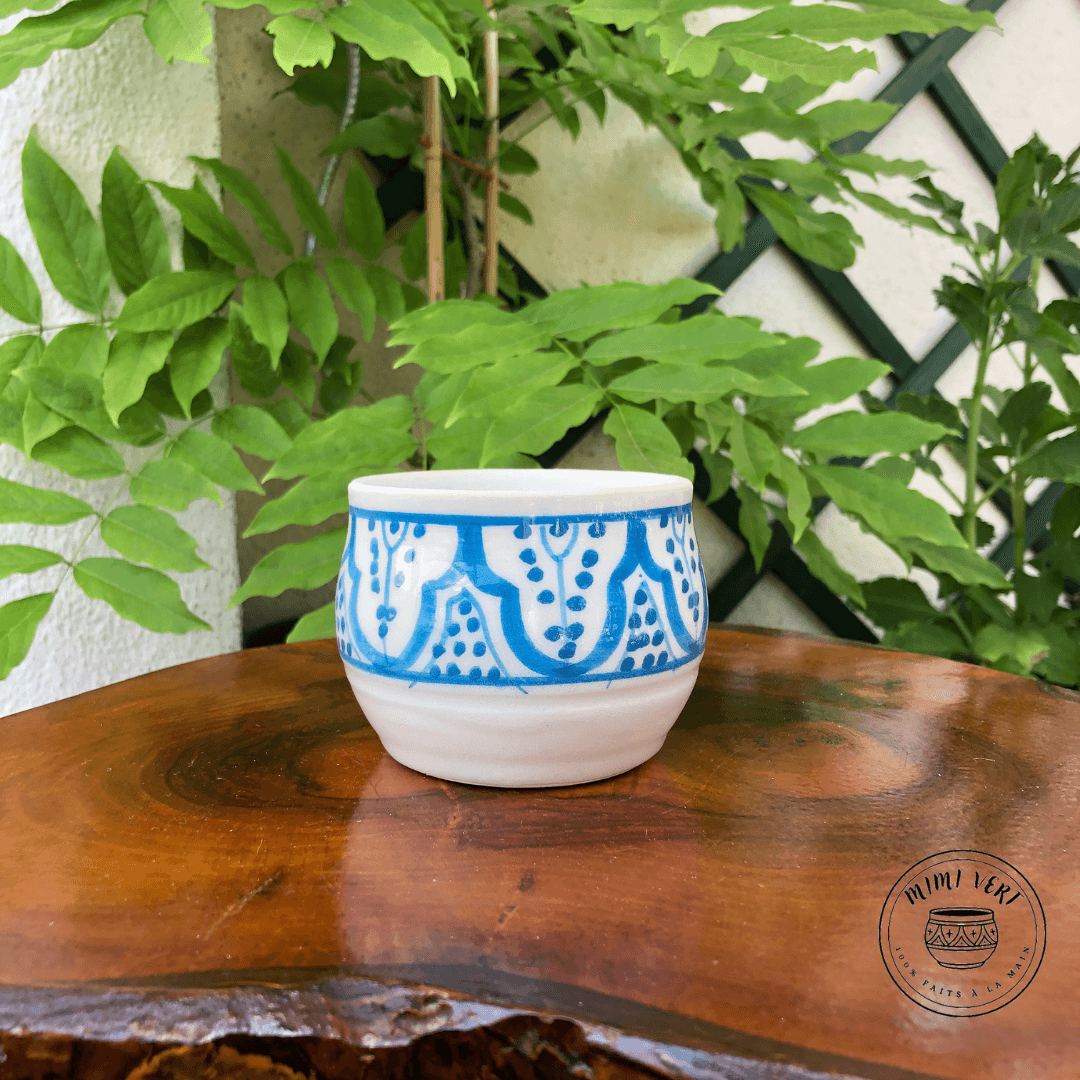 Pot en céramique fait à la main avec des motifs inspirés de l’artisanat marocain, blanc et bleu, contenant un cactus, posé sur une table en bois avec des plantes vertes en arrière-plan.