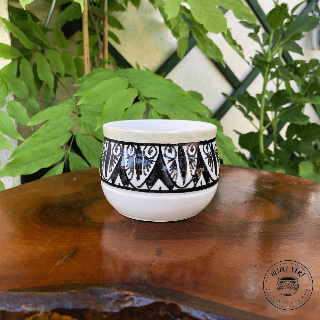 Pot en céramique fait à la main avec des motifs inspirés de l’artisanat marocain, noir et blanc, posé sur une table en bois avec des plantes vertes en arrière-plan.