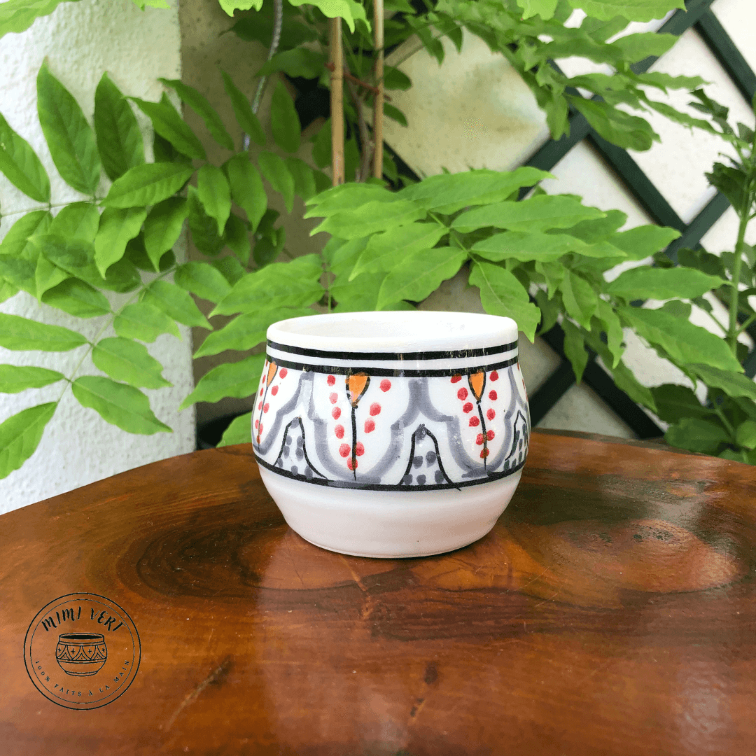 Pot en céramique fait à la main avec des motifs inspirés de l’artisanat marocain, blanc gris rouge jaune et noir, contenant un cactus, posé sur une table en bois avec des plantes vertes en arrière-plan.