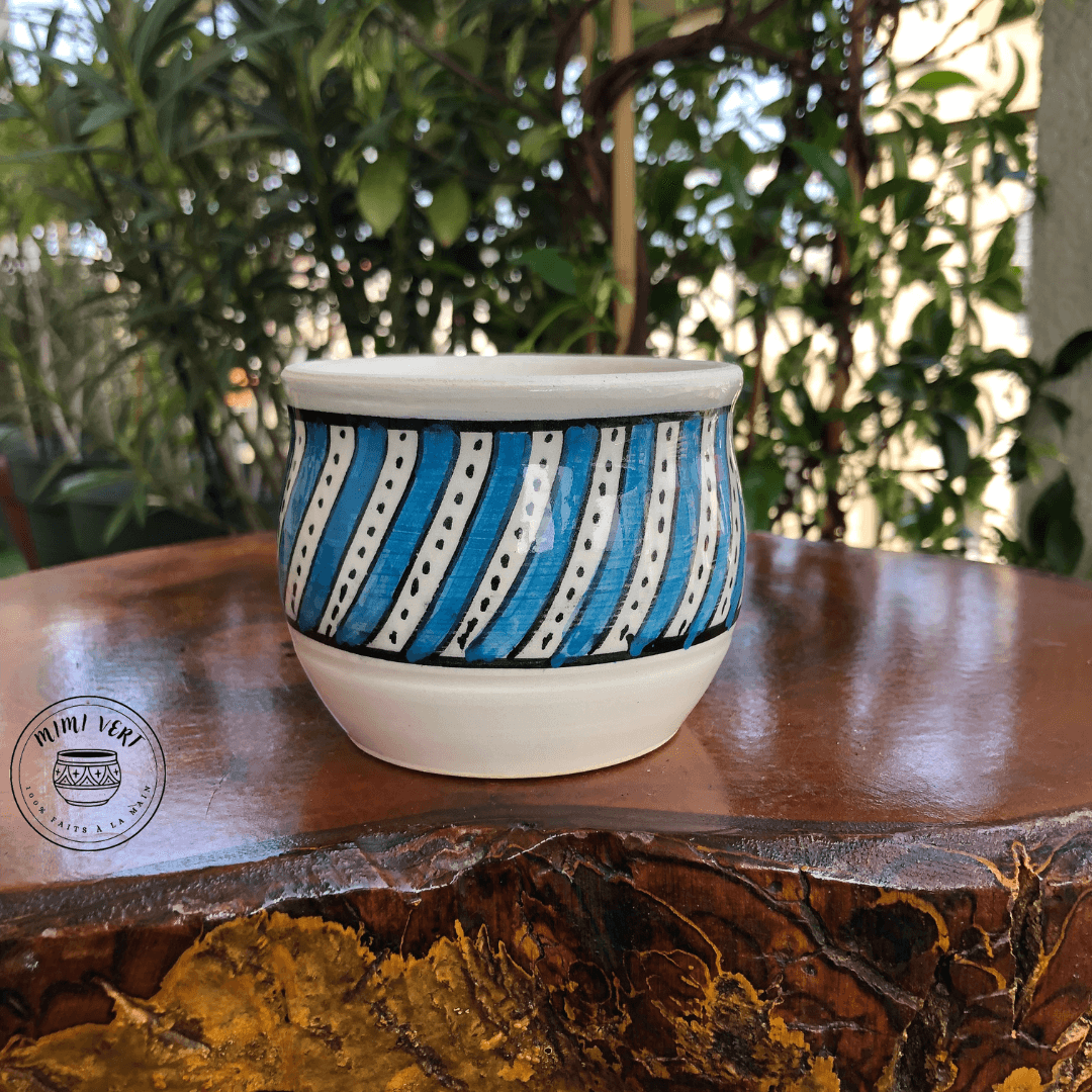 Pot en céramique fait à la main avec des motifs inspirés de l’artisanat marocain, bleu et blanc, posé sur une table en bois avec des plantes vertes en arrière-plan.