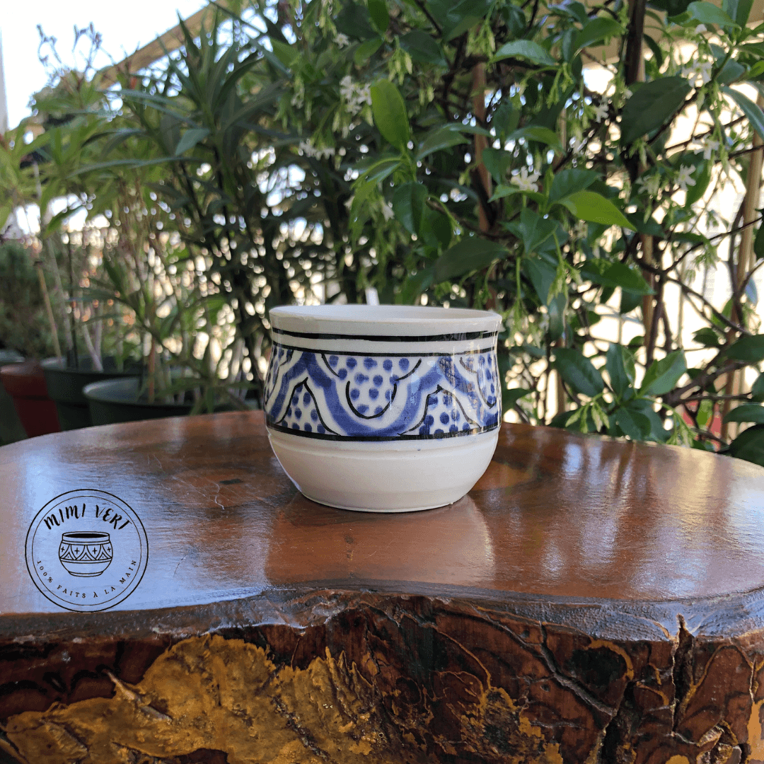 Pot en céramique fait à la main avec des motifs inspirés de l’artisanat marocain, bleu et blanc, posé sur une table en bois avec des plantes vertes en arrière-plan.