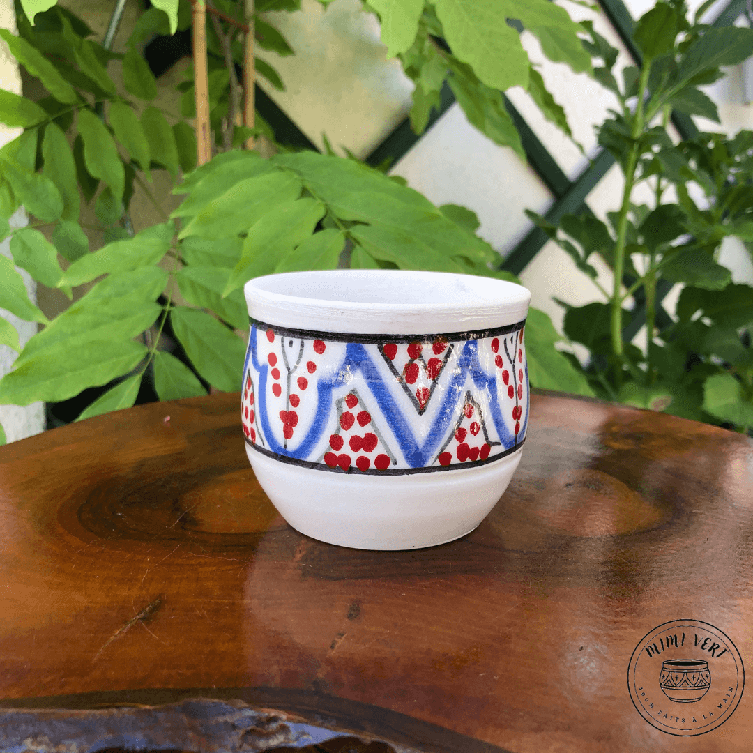Pot en céramique fait à la main avec des motifs inspirés de l’artisanat marocain, bleu noir et blanc et rouge, posé sur une table en bois avec des plantes vertes en arrière-plan.