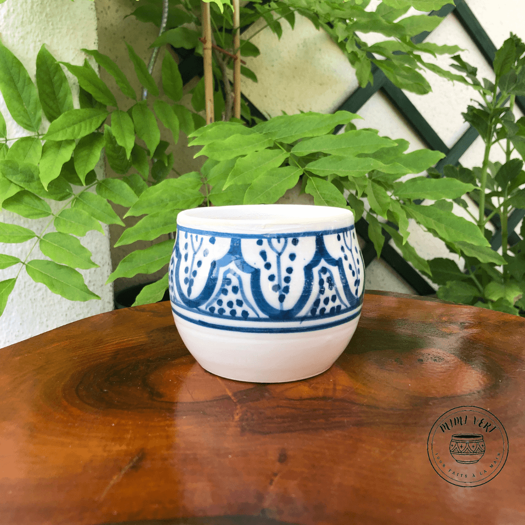 Pot en céramique fait à la main avec des motifs inspirés de l’artisanat marocain, blanc et bleu, contenant un cactus, posé sur une table en bois avec des plantes vertes en arrière-plan.
