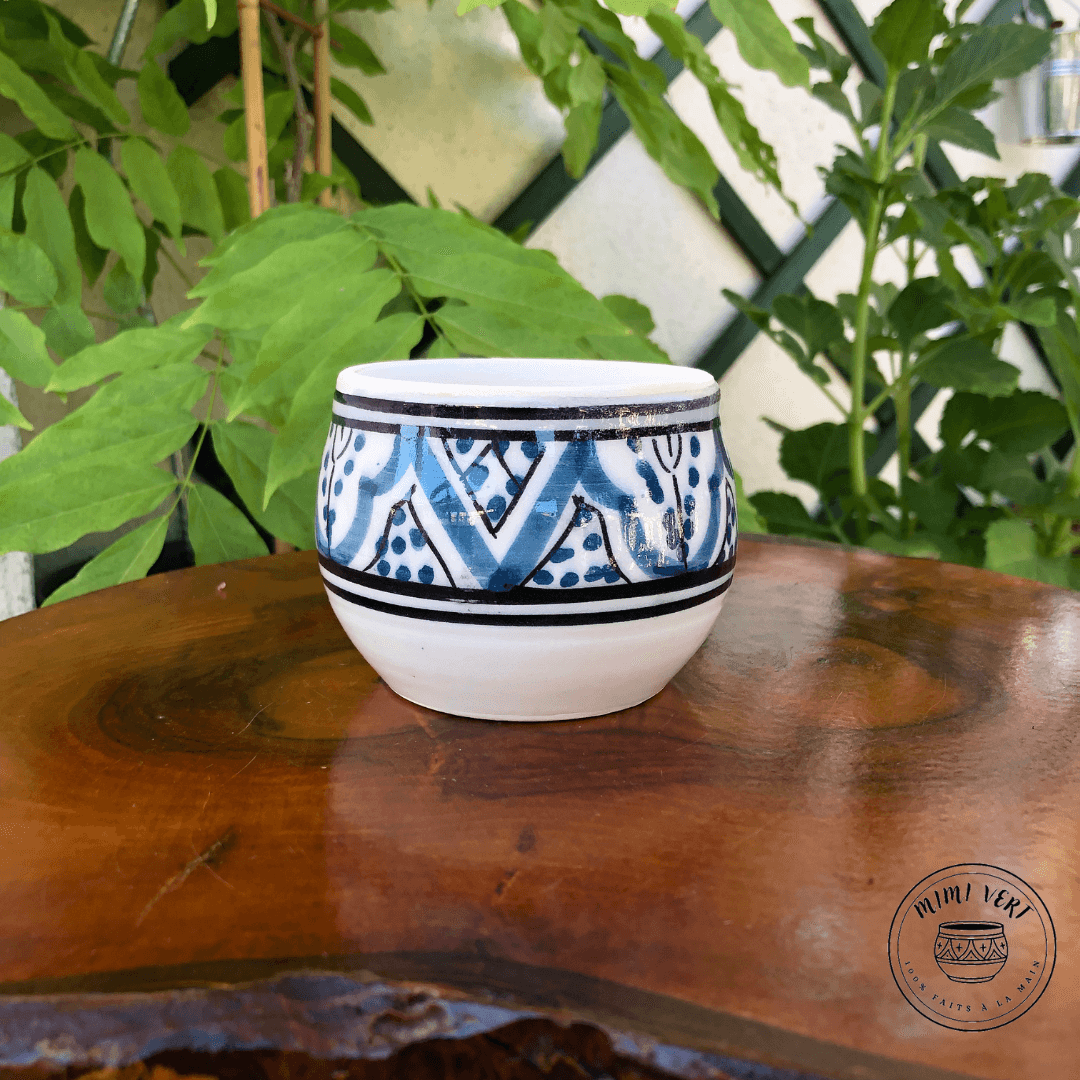 Pot en céramique fait à la main avec des motifs inspirés de l’artisanat marocain, bleu noir et blanc, posé sur une table en bois avec des plantes vertes en arrière-plan.