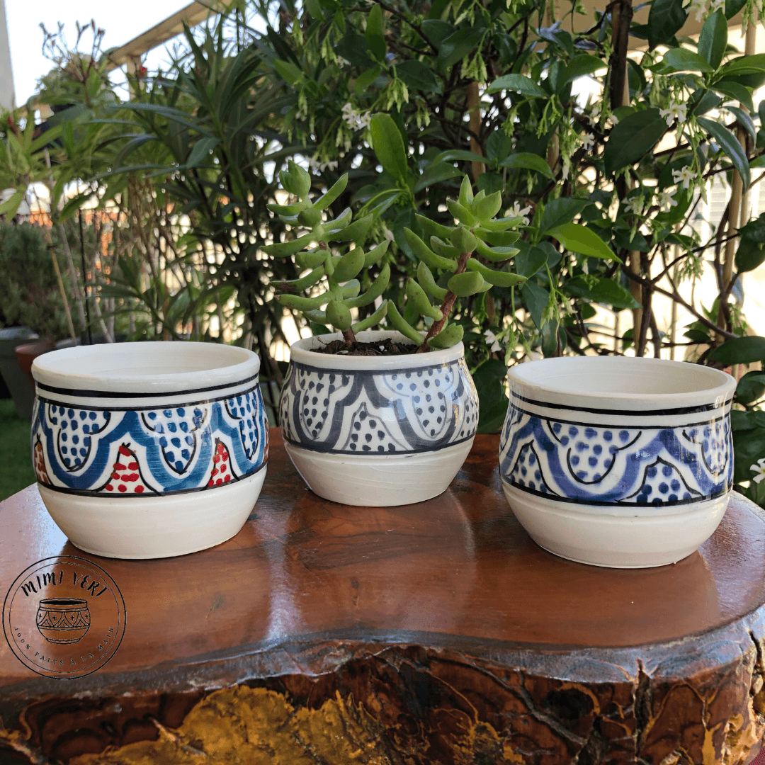 Trois pots en céramique faits à la main avec des motifs inspirés de l’artisanat marocain, posés sur une table en bois avec des plantes vertes en arrière-plan, dont un contenant une plante grasse.