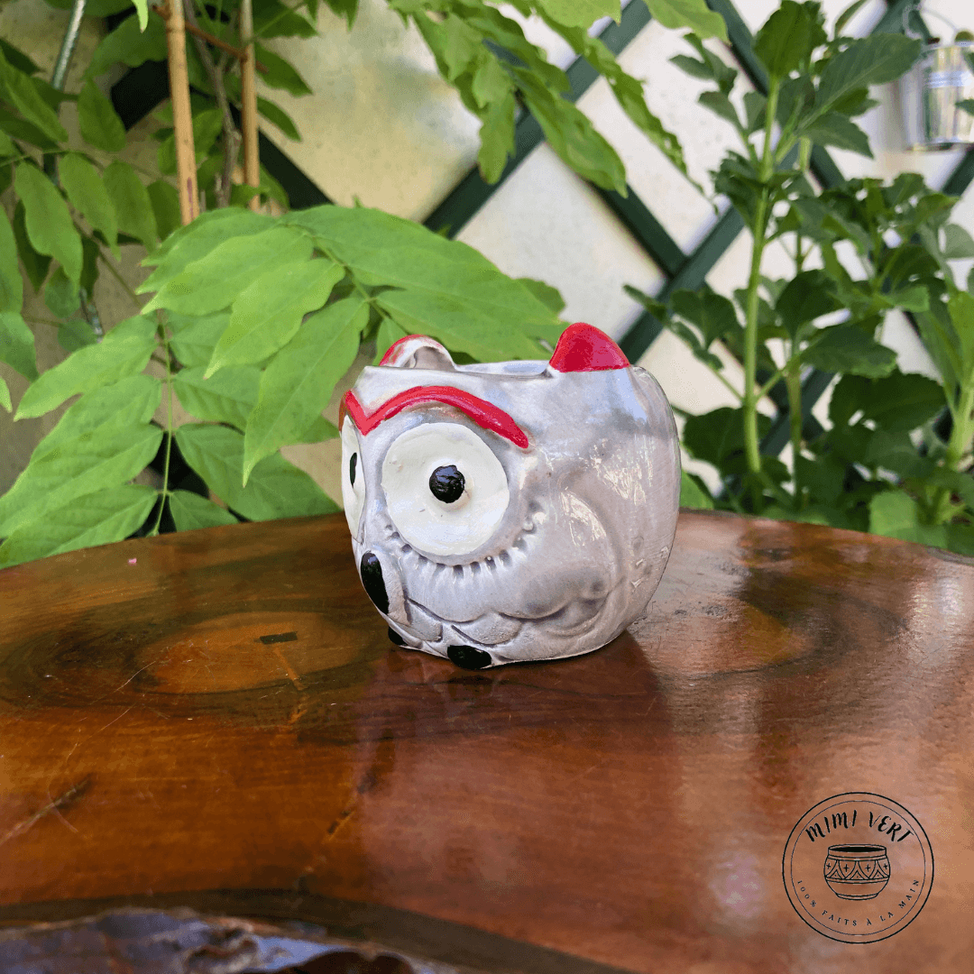 Petit pot en forme de hibou, gris clair, posé sur une table en bois avec des plantes vertes en arrière-plan.