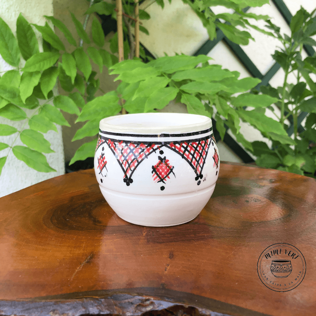 Pot en céramique fait à la main avec des motifs inspirés de l’artisanat marocain, blanc noir et rouge, contenant un cactus, posé sur une table en bois avec des plantes vertes en arrière-plan.