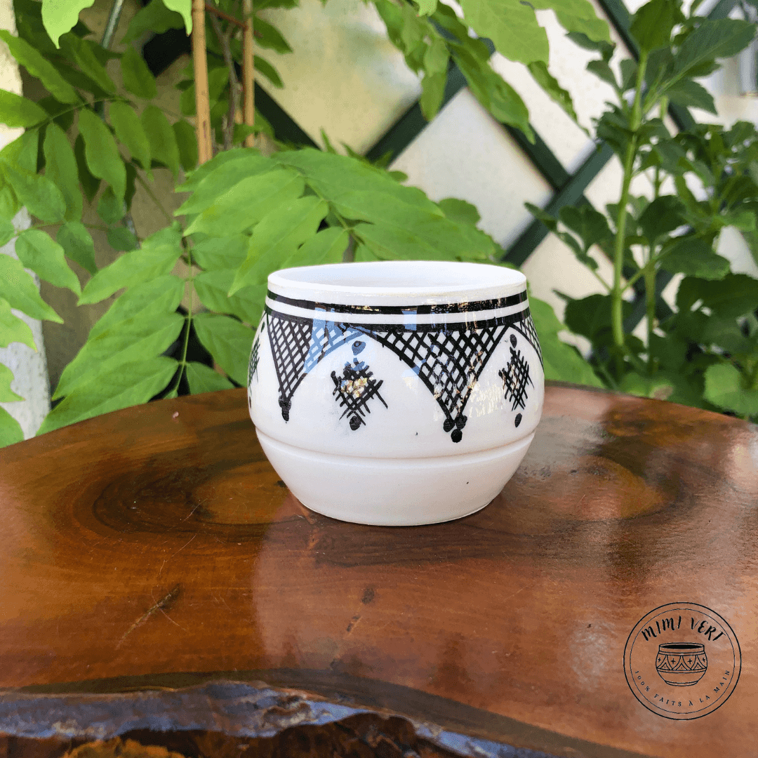 Pot en céramique fait à la main avec des motifs inspirés de l’artisanat marocain, noir et blanc, posé sur une table en bois avec des plantes vertes en arrière-plan.