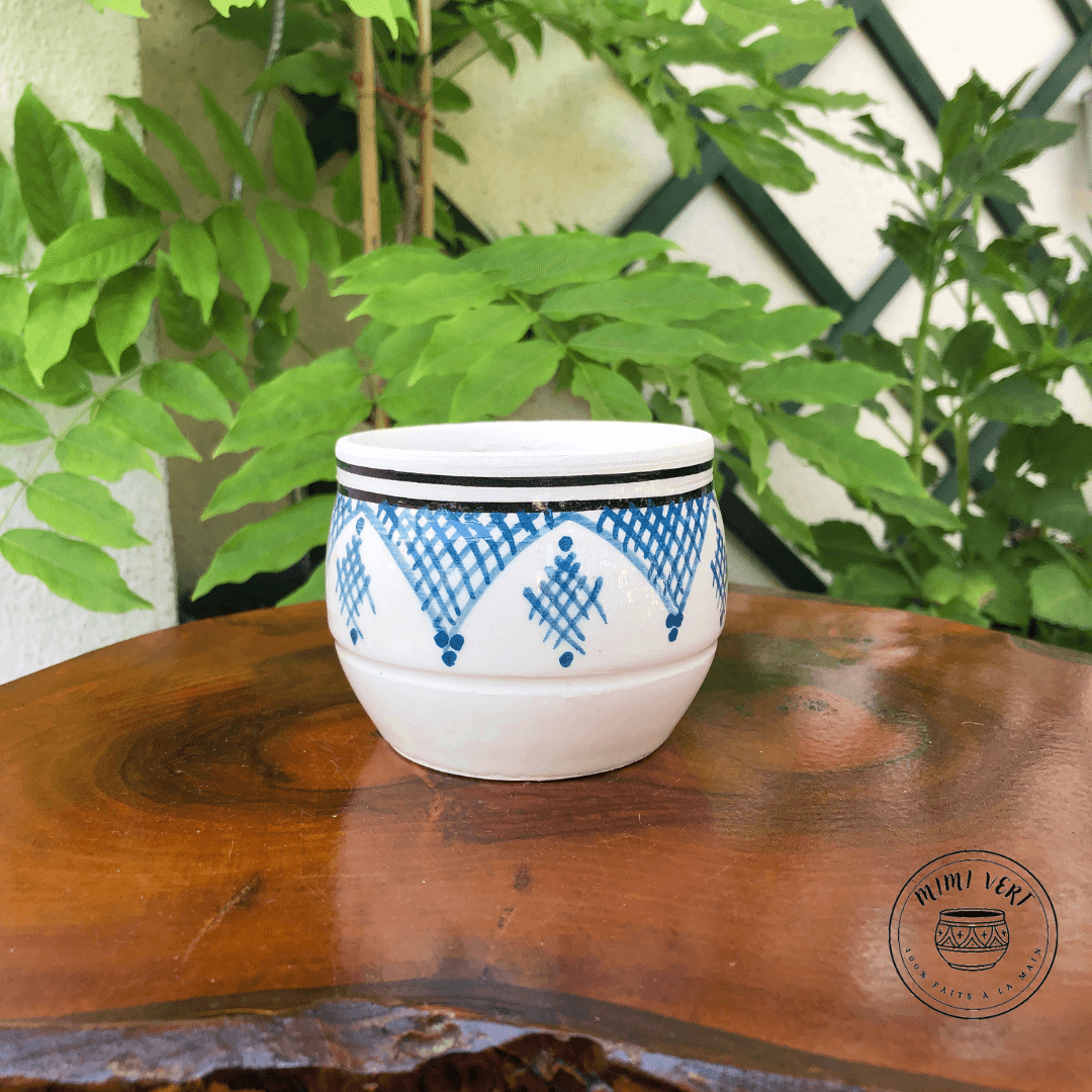 Pot en céramique fait à la main avec des motifs inspirés de l’artisanat marocain, blanc bleu et noir, contenant un cactus, posé sur une table en bois avec des plantes vertes en arrière-plan.