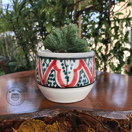 Pot en céramique fait à la main avec des motifs inspirés de l’artisanat marocain, blanc vert et rouge, contenant un cactus, posé sur une table en bois avec des plantes vertes en arrière-plan.